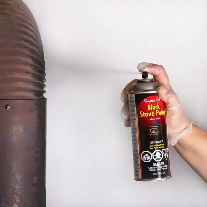 Hand holding Imperial Black Stove Paint aerosol can near stove pipe, designed for temperatures up to 700°C, ideal for stoves and barbecues.