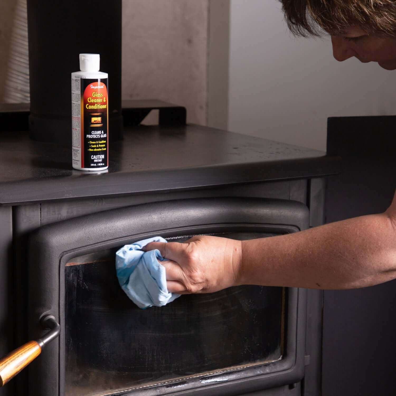 Cleaning wood stove glass with Imperial Glass Cleaner And Conditioner from Cabin Depot, removing soot and creosote for a clear view.