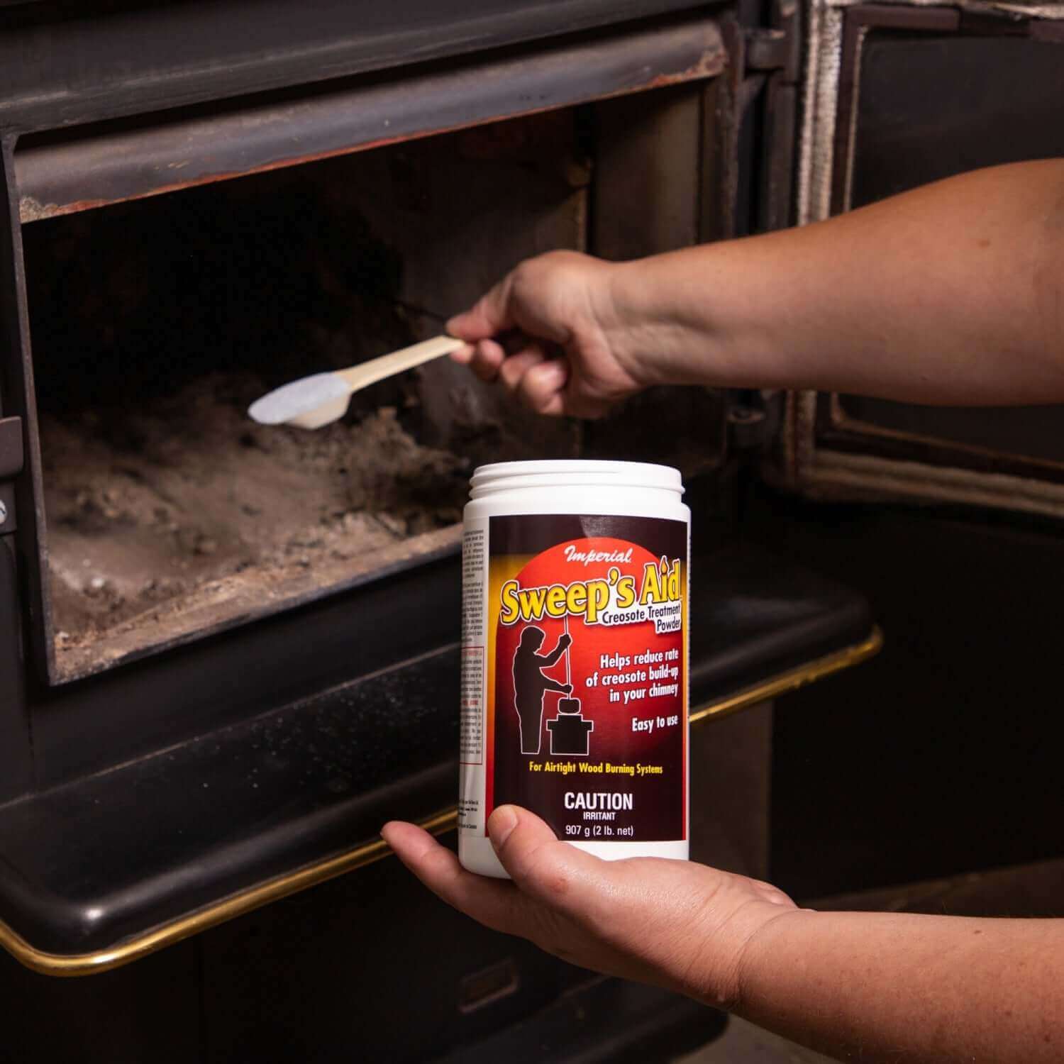 Person applying Imperial Sweep's Aid Stove Cleaner in wood stove for improved performance at Cabin Depot.