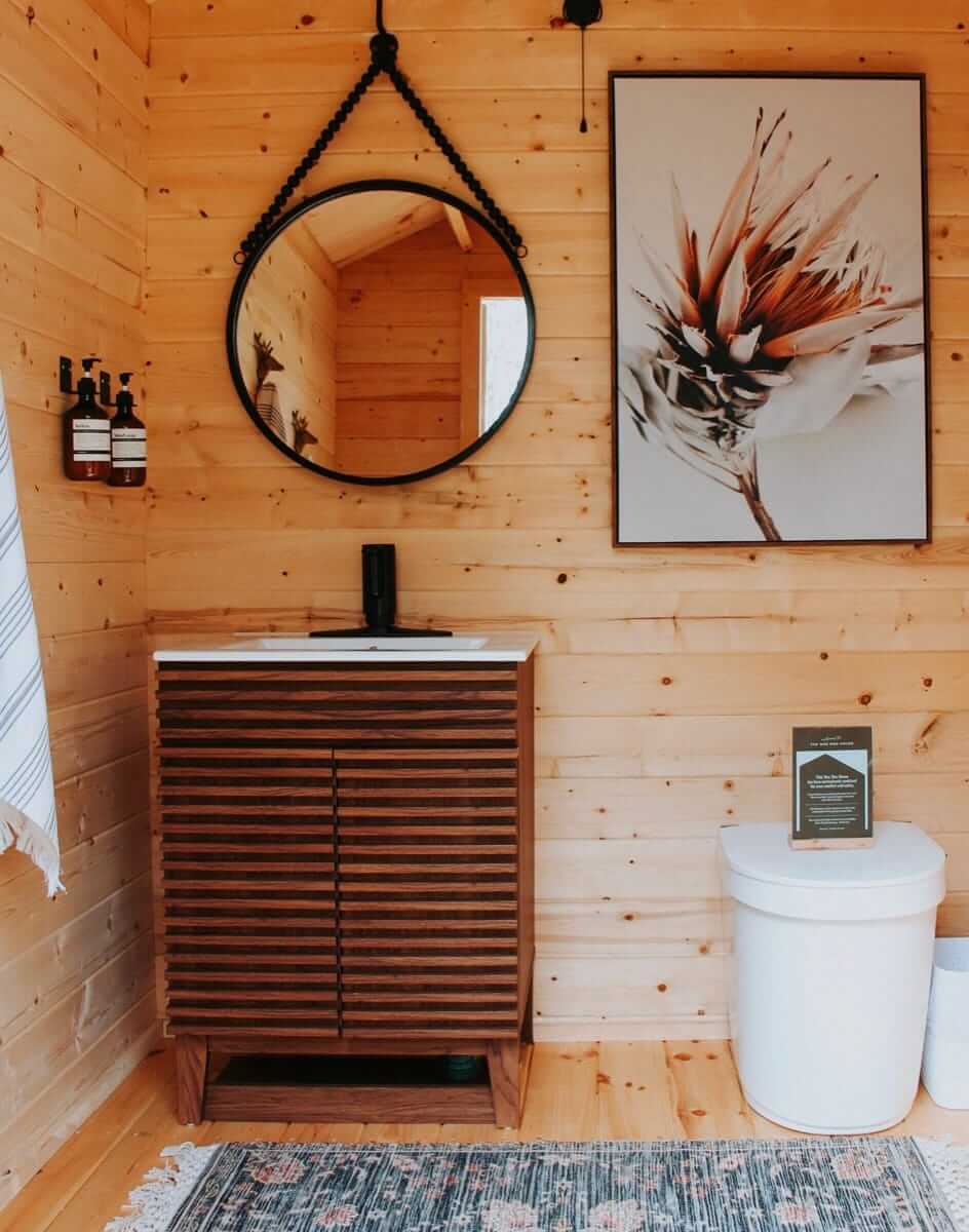 Interior of Loo Loo House Log Outhouse showing modern design with wooden walls, a round mirror, and a compact toilet. Cabin Depot inspired.