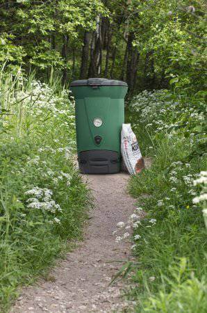 Green Biolan 220 ECO Composter on garden path surrounded by lush greenery and wildflowers.