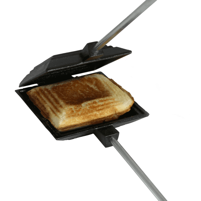 Cast iron camp cooker toasting a sandwich over an open fire, featuring a durable hinge and wooden handles for easy transport.