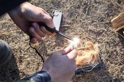 Hands using Coghlan's Fire-In-A-Box kit to ignite tinder with flint and magnesium for outdoor campfire.