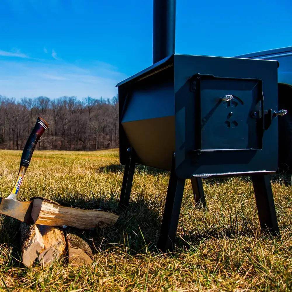 Englander Grizzly Kit Wood Stove on grass with log and axe, available at Cabin Depot for outdoor and camping use. Durable 12-gauge steel.