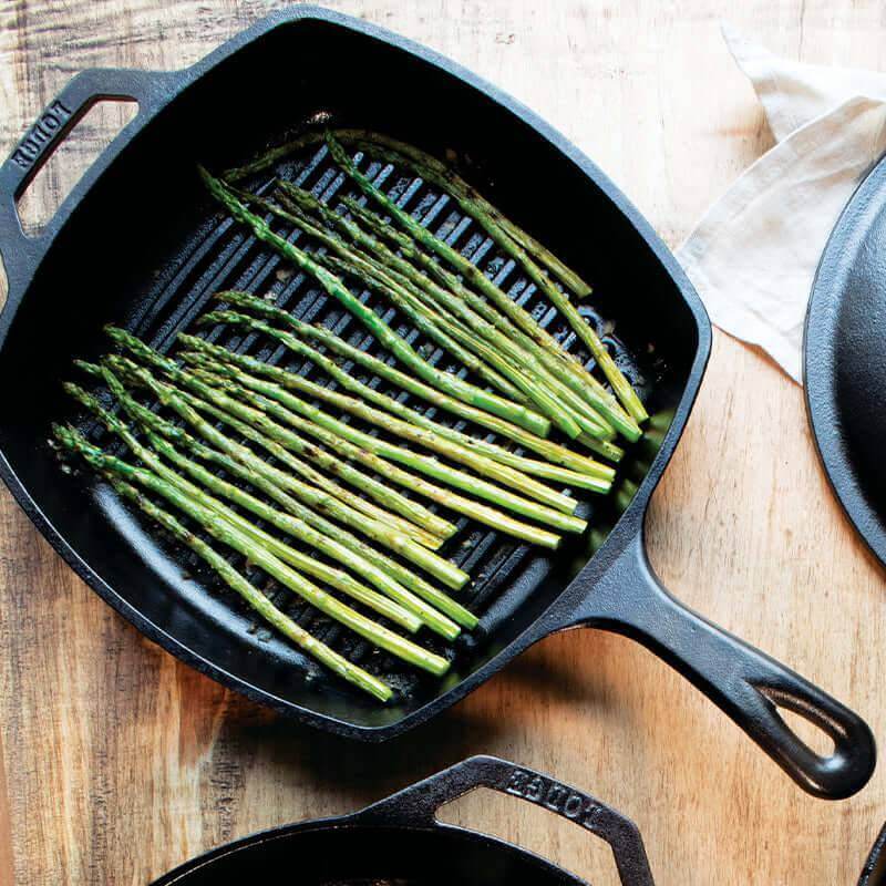 Lodge 10.5" Cast Iron Square Grill Pan from Cabin Depot with grilled asparagus, seasoned for unbeatable heat retention.