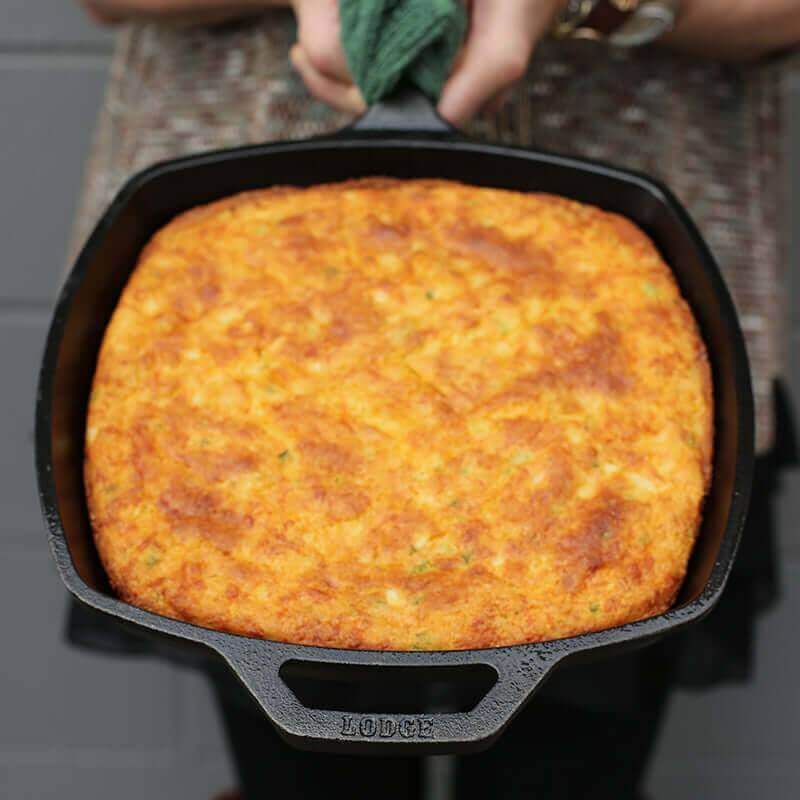 Person holding Lodge 10.5" cast iron square skillet with cornbread, seasoned for cooking, available at Cabin Depot.