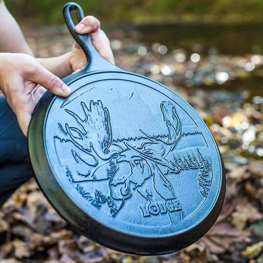 Lodge 10.5" Wildlife Series Cast Iron Griddle with Moose Design, held outdoors, showcasing seasoned cooking surface.