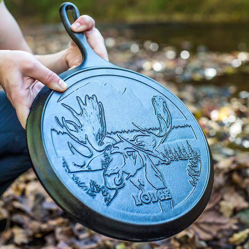 Person holding Lodge 10.5 inch Cast Iron Griddle with Moose design near a stream, available at Cabin Depot.