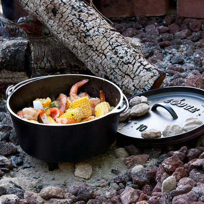 Lodge 10" 5 Quart Camp Dutch Oven cooking seafood and corn over campfire coals, with lid on rocky ground.