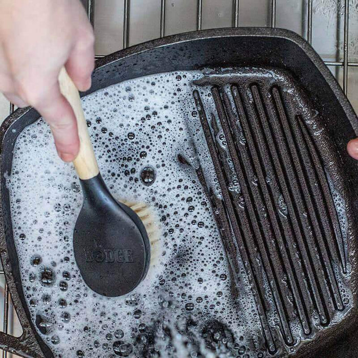 Lodge 10" Scrub Brush cleaning cast iron pan with soapy water and nylon bristles for gentle scrubbing on enameled surfaces.