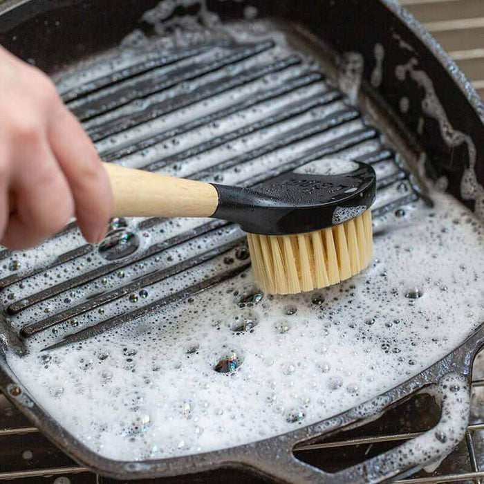 Scrubbing cast iron grill pan with Lodge 10" scrub brush, featuring a comfort handle and gentle nylon bristles with soap suds.