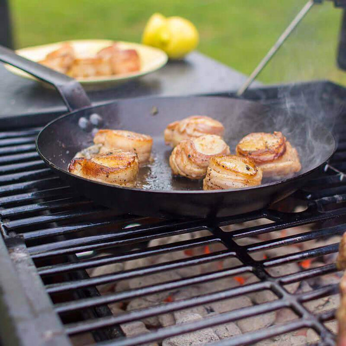 Grilling meat in Lodge 10" seasoned steel skillet over open flame, perfect for home chefs and camp cooks, with easy-release finish.