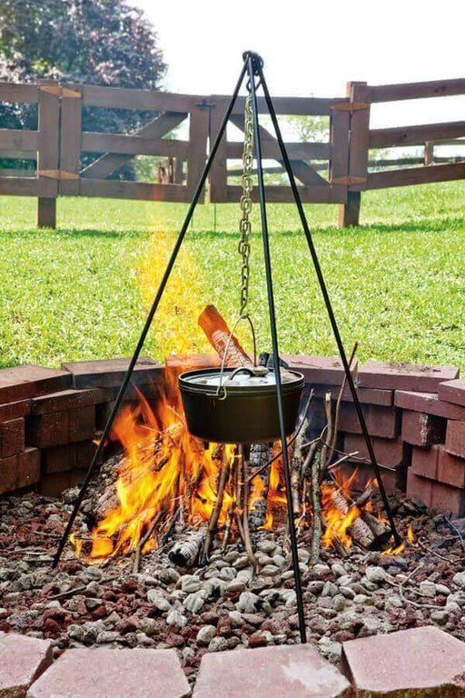 Campfire cooking with Lodge 60" tripod and dutch oven over open flames, featuring solid steel construction and galvanized chain.