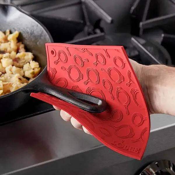 Red Lodge silicone trivet with skillet pattern protecting hand while holding a hot pan near stovetop.