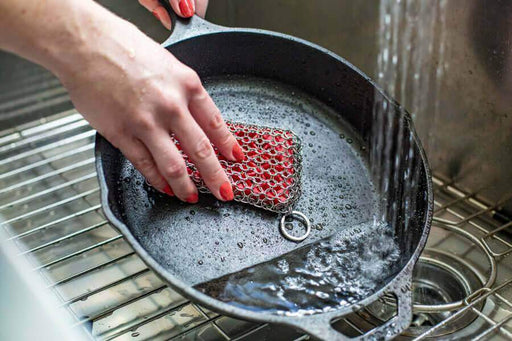 Hand cleaning cast iron pan with Lodge Chainmail Scrubbing Pad under running water in kitchen sink.