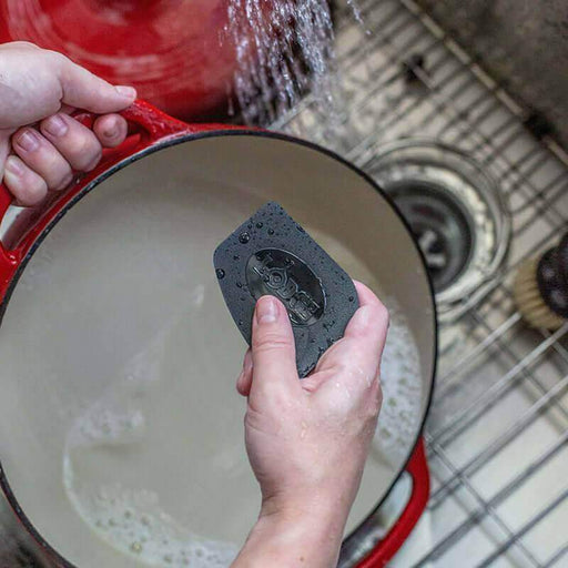 Person using Lodge pan scraper to clean red pot under running water in kitchen sink.