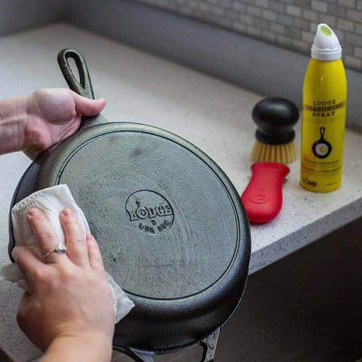 Cleaning cast iron skillet with Lodge Seasoned Cast Iron Care Kit, featuring seasoning spray, scrub brush, and handle holder on counter.