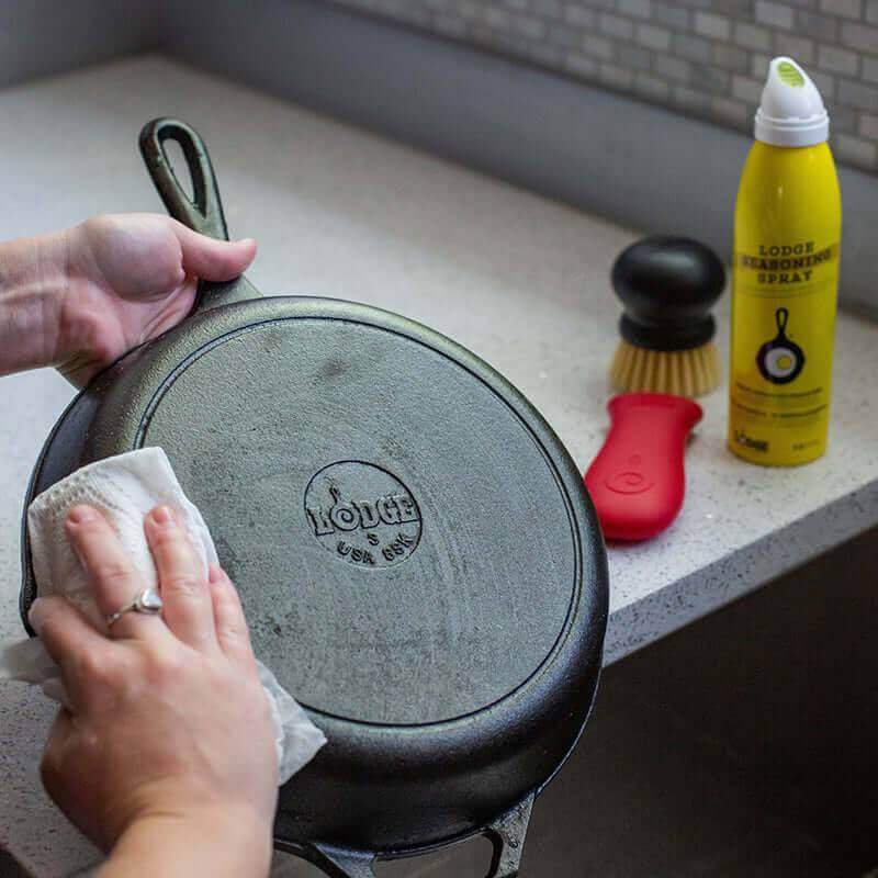 Person cleaning cast iron skillet with Lodge Seasoned Cast Iron Care Kit, featuring oil spray, scrub brush, scraper. Available at Cabin Depot.