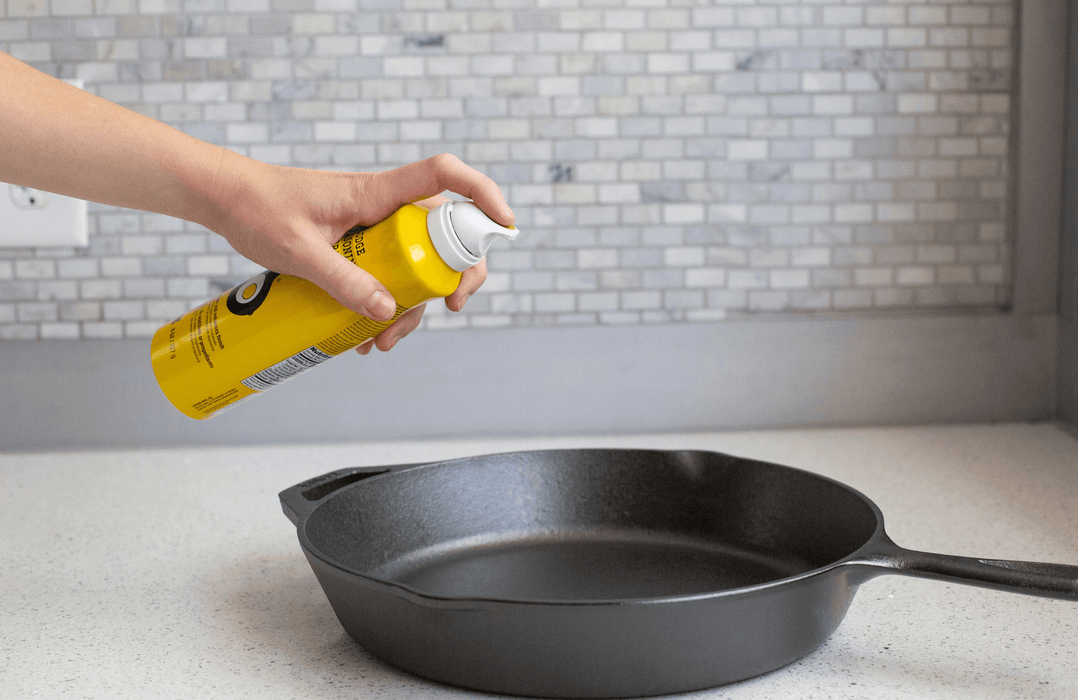 Person using Lodge Seasoning Spray on cast iron skillet for protection and enhanced finish on kitchen counter.