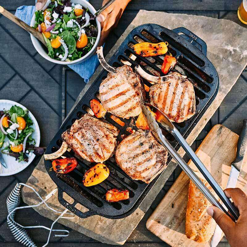 Alt Tag: Lodge Sportsman's Pro Cast Iron Grill from Cabin Depot with sizzling BBQ chops and veggies, ideal for tailgates and cookouts.