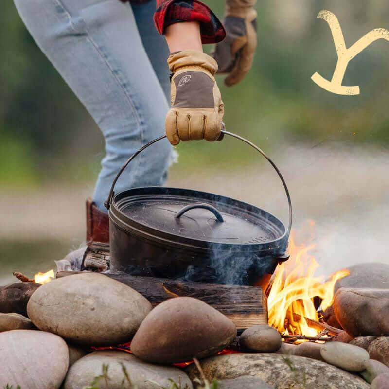 Lodge Yellowstone 12'' Cast Iron Camp Dutch Oven over fire, featuring Power Y lid, perfect for camping. Available at Cabin Depot.