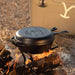 Lodge Yellowstone cast iron combo cooker with bucking bronco design on lid, sitting on campfire, perfect for outdoor cooking.