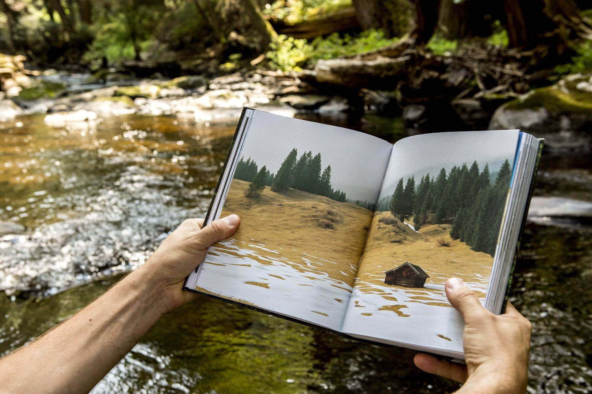 Person reading Cabin Porn book by a stream, showcasing a rural landscape illustration, highlighting Cabin Depot's nature retreat inspiration.