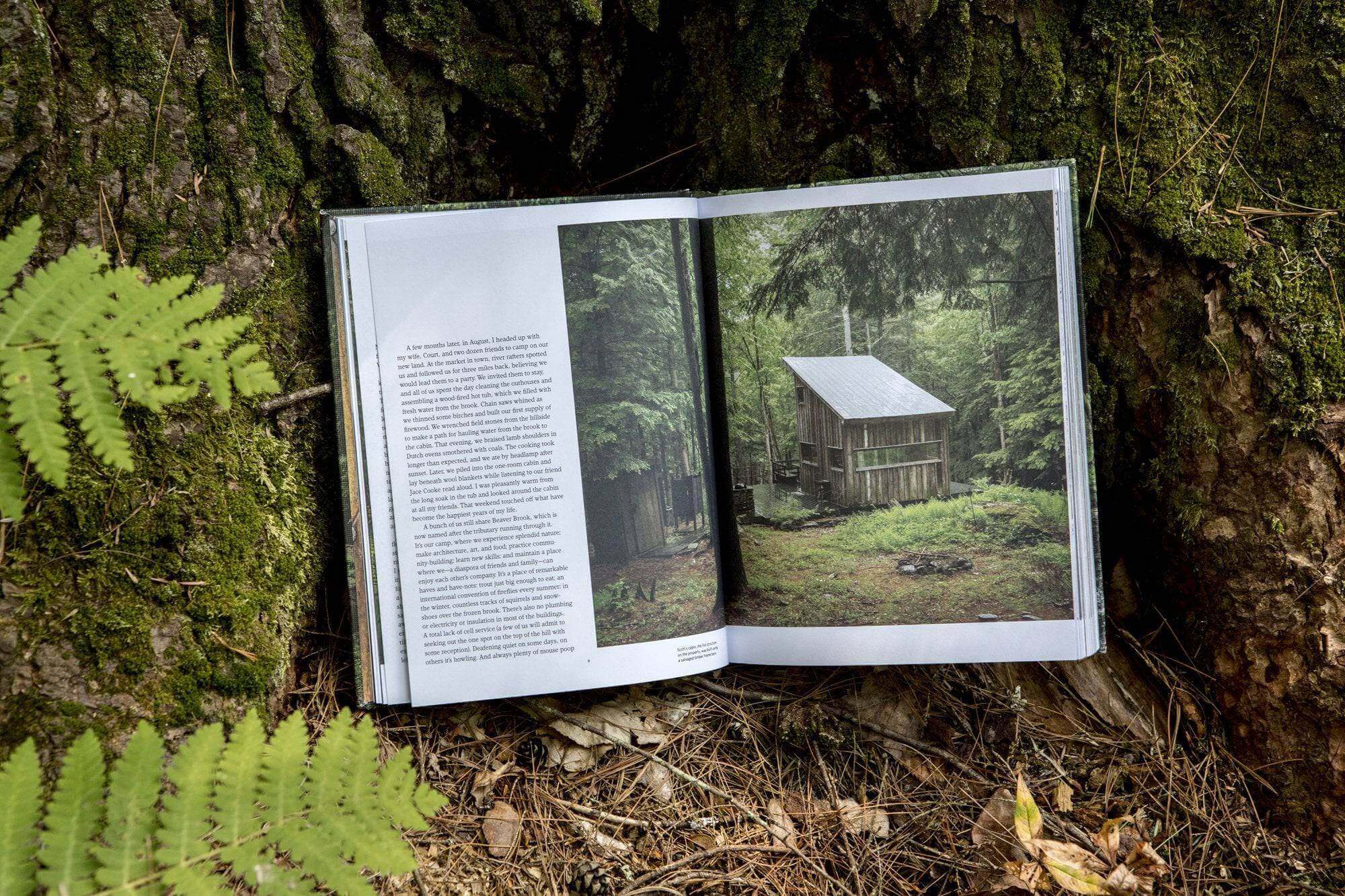 Cabin Porn book open to a page with a rustic cabin photo, nestled by a tree in a forest, epitomizing rural escapes. Cabin Depot inspiration.