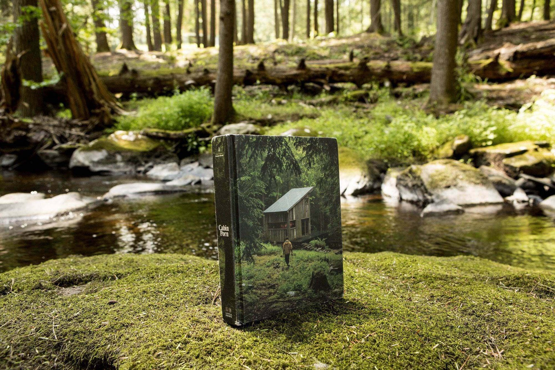 Cabin Porn book displayed on mossy rock by forest stream, showcasing rural retreat imagery from Cabin Depot.