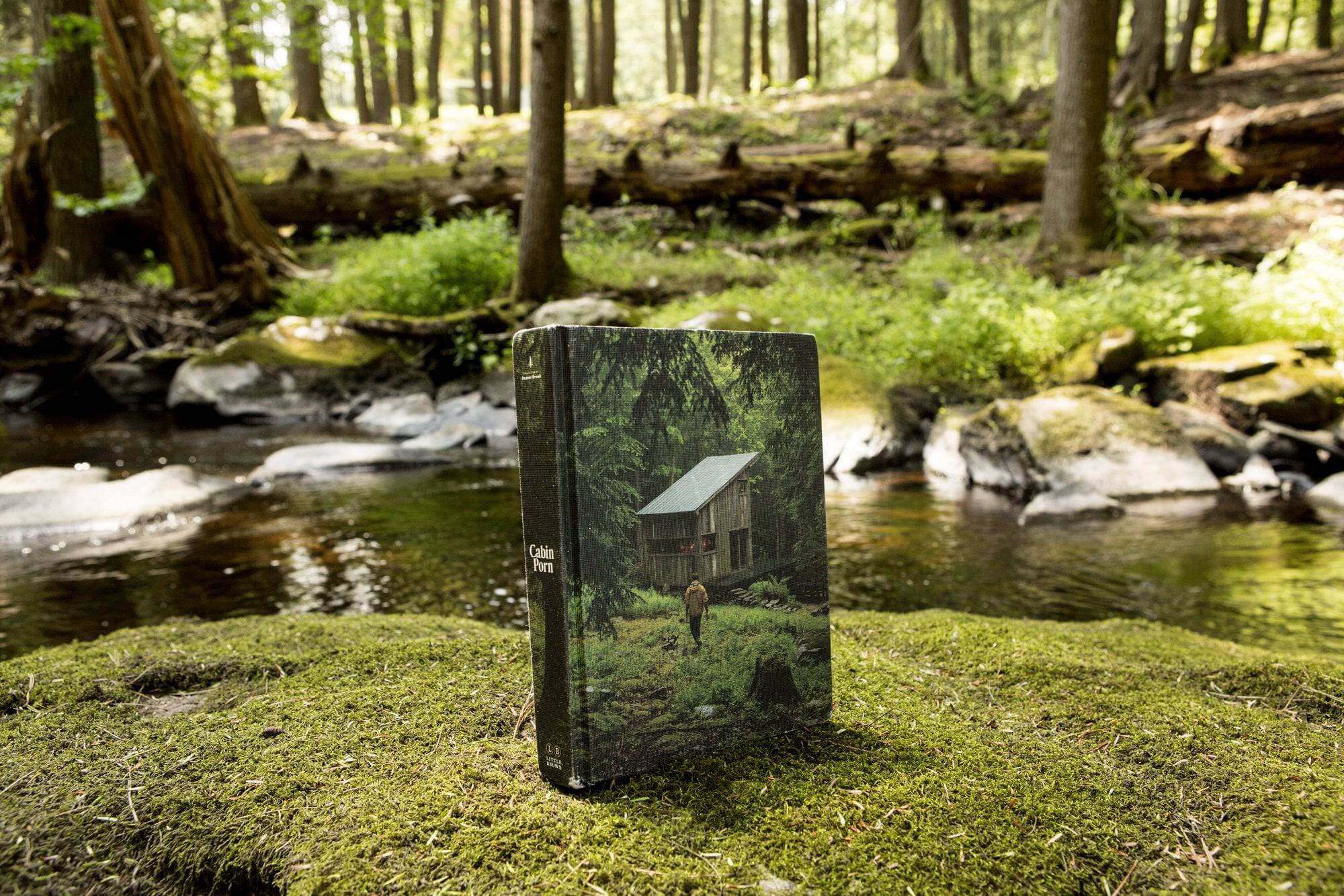 Cabin Porn book displayed on mossy rock by forest stream, showcasing rural retreat imagery from Cabin Depot.
