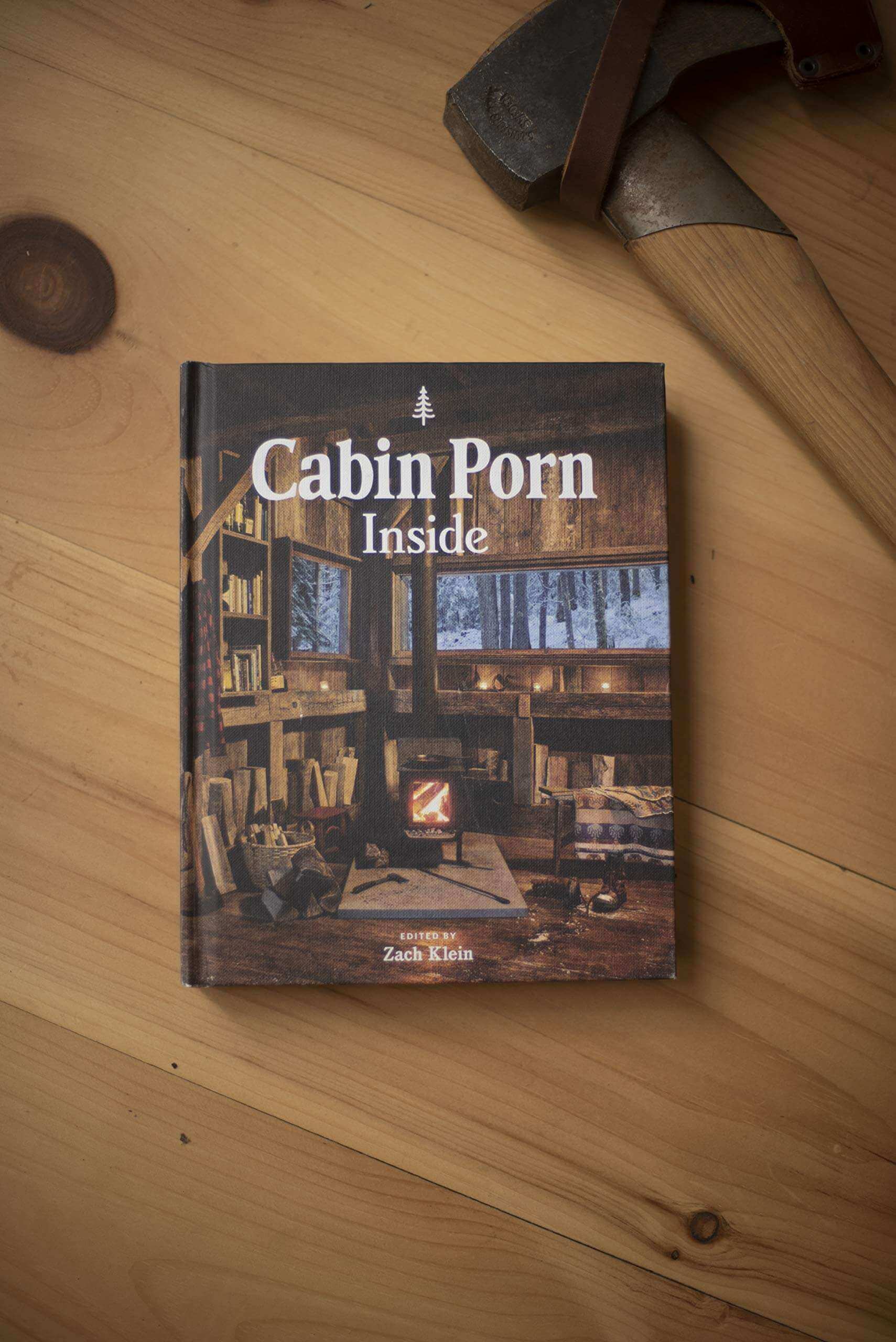 Cabin Porn Inside book on wooden table near axe, available at Cabin Depot, inspiring homebuilding and cabin design.