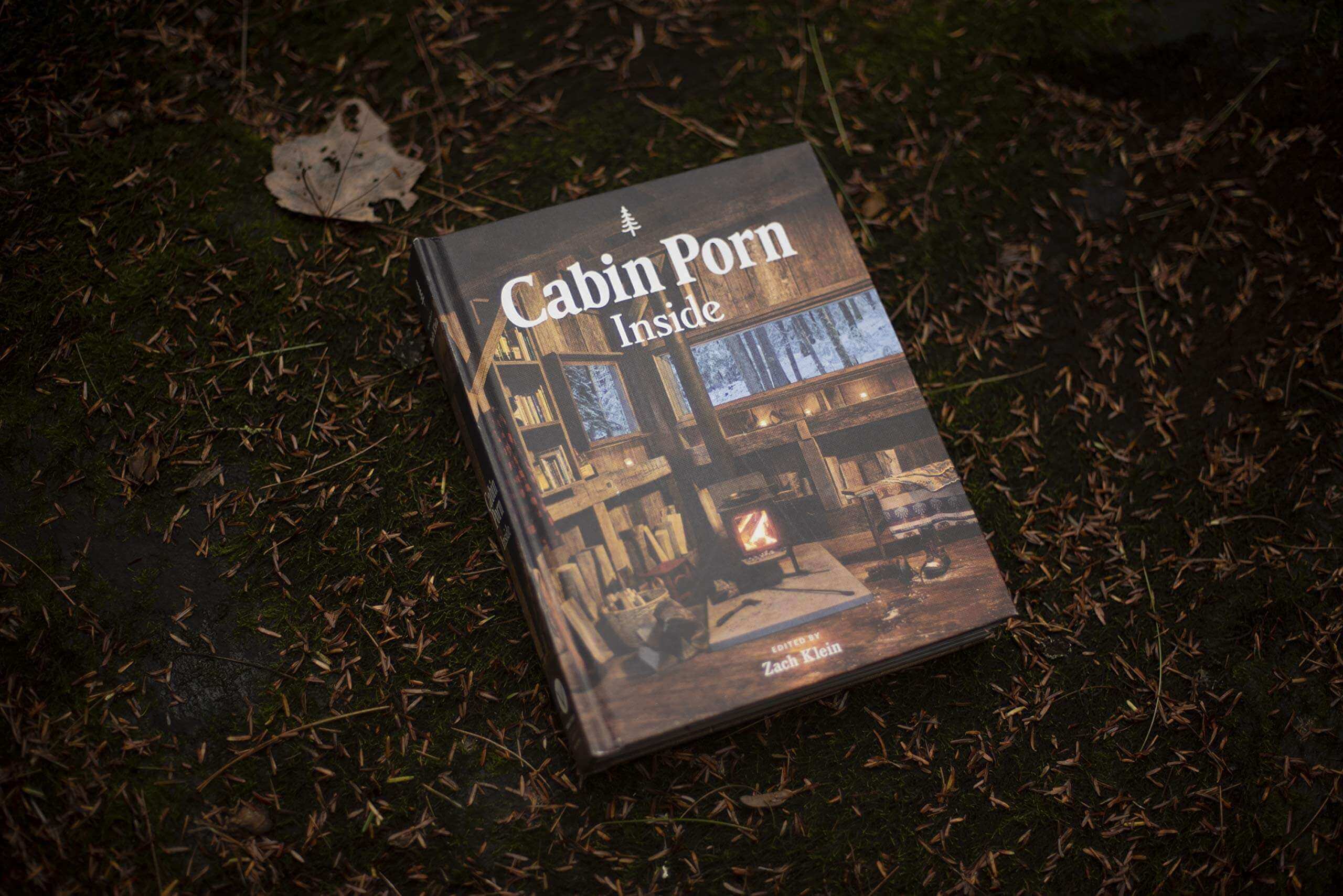 Cabin Porn Inside book cover on forest floor, featuring cozy cabin interior, available at Cabin Depot for homebuilding inspiration.