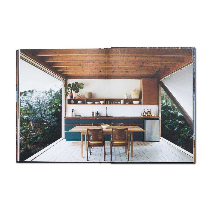 Open book showing a tranquil cabin interior with a wooden dining table and lush greenery outside through large windows.