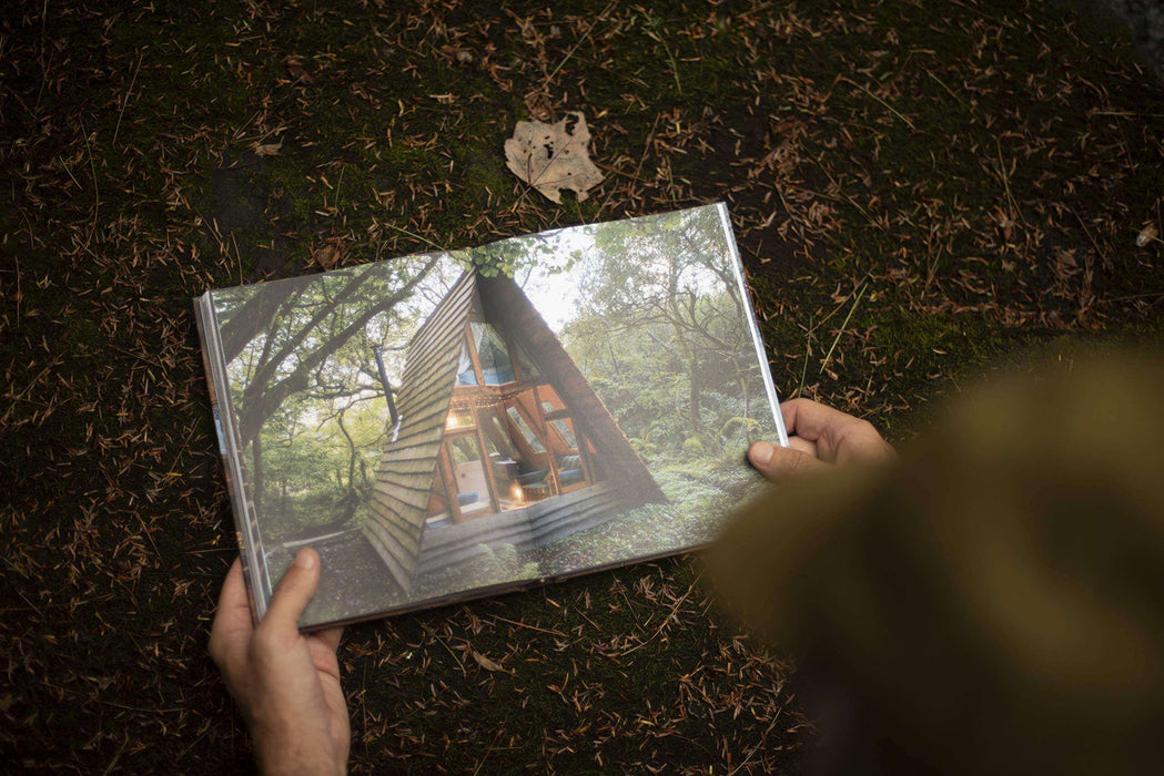 Person holding Cabin Porn Inside Book open to a photo of a serene woodland cabin.