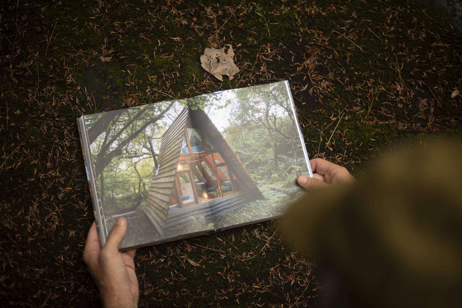 Person reading Cabin Porn Inside Book featuring a serene forest cabin scene, available at Cabin Depot for aspiring home builders.